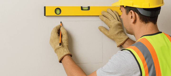 Construction worker using a level to mark the precise cutout location for a drywall access door on a wall.